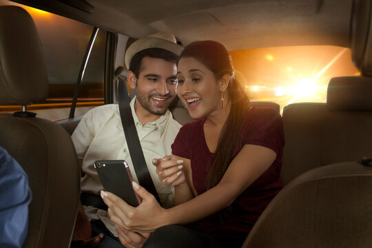 Happy young couple using mobile phone sitting on the back seat of car