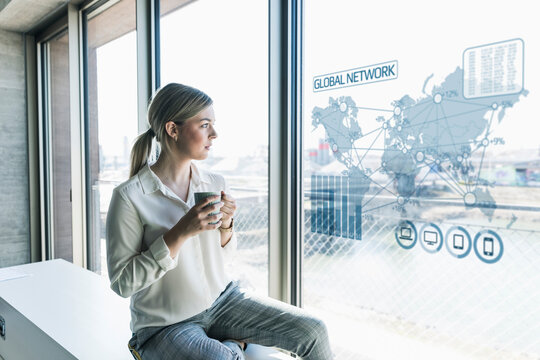 Young businesswoman looking at virtual world map at window pane in office