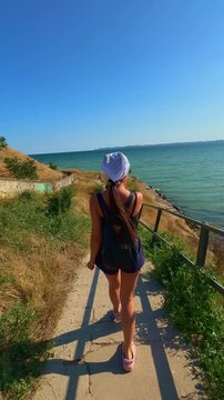 Rear view of a young woman with a backpack and hat walking down a sunny coastal path towards the blue sea. Beautiful summer landscape. Ideal for travel vlogs, lifestyle commercials, and vacation ads.