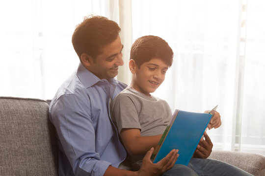 Father and son reading book sitting on sofa
