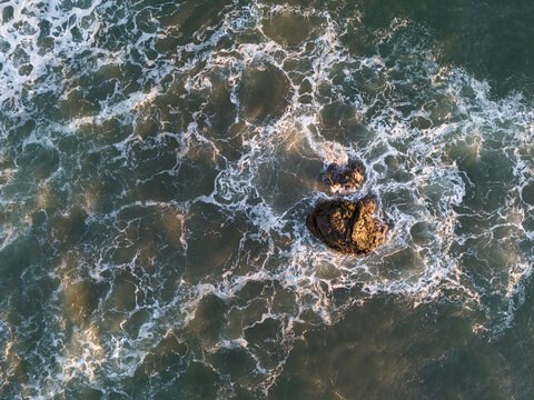 Aerial view of ocean waves crashing against dark rocks in the sea with white foam patterns and golden sunlight reflecting on the water in Huehuete, Carazo, Nicaragua.