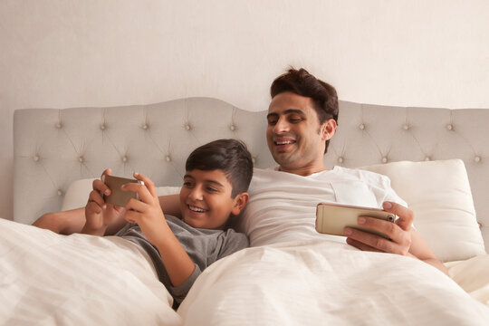 Happy father looking at son playing games on smart phone in bed