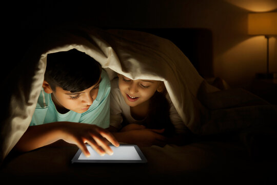 Brother and sister sharing digital tablet under blanket at night
