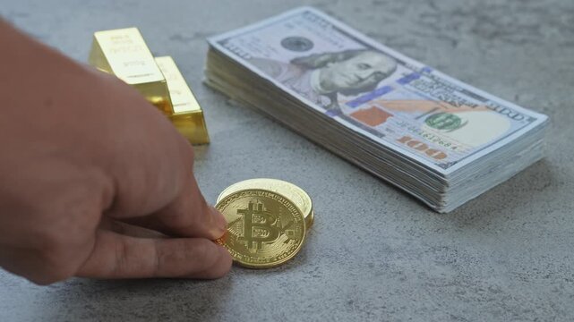 Hand arranges gold bitcoin coins next to gold bars and a stack of US dollar bills, representing financial assets