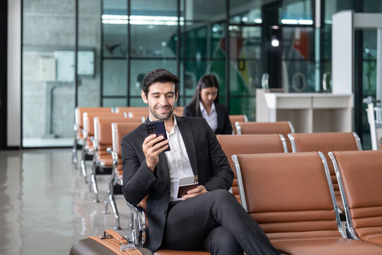 young male executive in suit holding passport with boarding pass, sitting in modern airport terminal checking time on a smartphone,preparing for corporate business trip or international flight