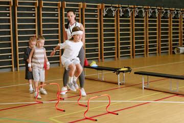 Obraz premium Caucasian female trainer supervising children jumping over hurdles in school gym. Image suitable for youth sports programs, physical education marketing, and healthy lifestyle campaigns