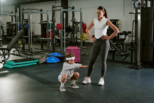 Caucasian female trainer guiding Caucasian child doing squats in modern gym. Image suitable for promoting youth fitness programs, personal training services, and healthy lifestyle habits