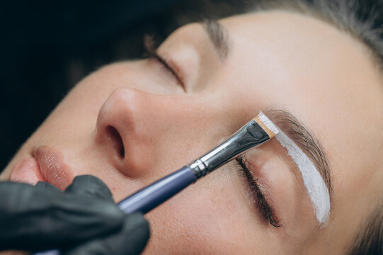 Permanent makeup artist applies white mapping paste to outline eyebrow shape before a cosmetic tattoo procedure in a professional beauty studio.