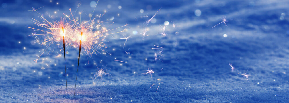two burning sparklers with flying stars isolated in snow, party scenery on blurred winter background with copy space, greeting card for december holiday season