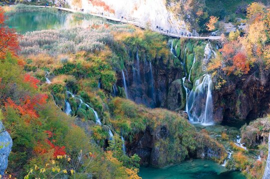 A gorgeous high-angle vista captures the majestic Milanovac Waterfall pouring into the turquoise pools of Plitvice Lakes, bordered by a vibrant autumn forest and a crowded cliffside boardwalk