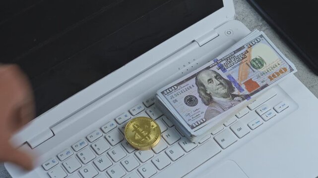 Hand places gold bitcoin next to stack of US dollars on laptop keyboard for online financial trading concept
