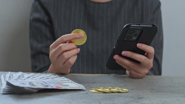 Woman holds gold bitcoin while checking smartphone with stacks of cash and crypto coins on desk