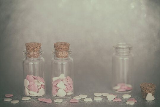 Two glass jars filled with Assorted pink and white Heart shaped sprinkles and an open glass jar with sprinkles around it on a table