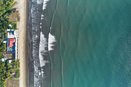 Aerial view of Quizala beach with gentle waves breaking on the dark sand, surrounded by lush vegetation and coastal buildings in San Rafael del Sur, Managua, Nicaragua.