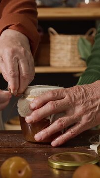 Vertical video of senior couple hands tying twine around cloth lid of homemade jam jar. Clip is ideal for organic food brands and local market promotions