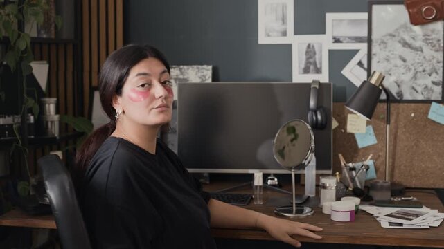Portrait of young adult woman applying pink under-eye patches at desk, turning to look at camera and smiling. Footage ideal for skincare, self-care, and remote work wellness campaigns