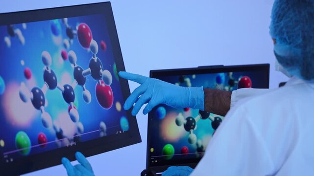 Asian female lab technician in protective mask pointing at molecular structure display on interactive monitor screen during biochemistry analysis inside laboratory