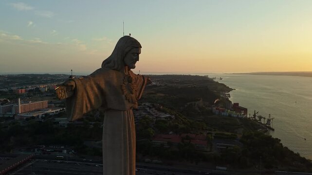 Close aerial view of the Cristo Rei monument illuminated by warm sunset light above Almada, Portugal, with the Tagus estuary and urban landscape in the background.