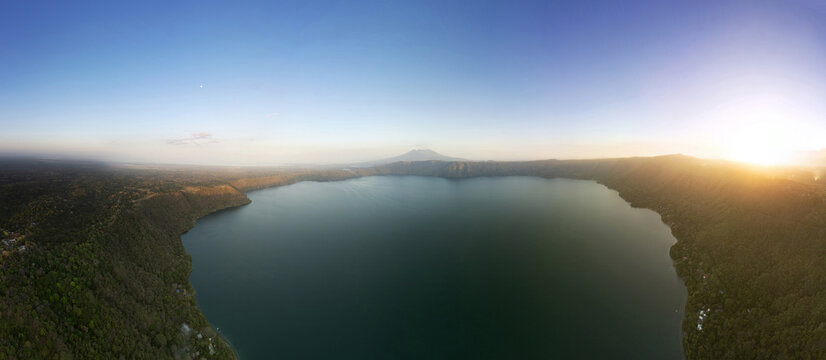 Aerial view of Apoyo Lagoon surrounded by lush tropical forests and volcanic hills under a clear sky at sunset, Masaya, Masaya, Nicaragua.