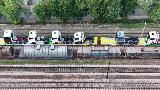 New trucks being transported on railway platforms, showcasing a continuous flow of vehicles on flatbed cars, with surrounding greenery and railway tracks visible in the background. Aerial view