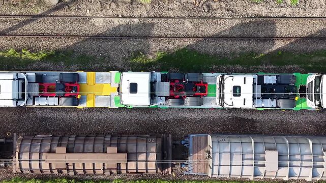 New trucks being transported on railway platforms, showcasing a continuous flow of vehicles on flatbed cars, with surrounding greenery and railway tracks visible in the background. Aerial view