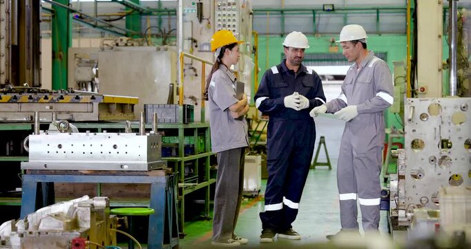 Three adult factory workers examine production document together, coordinating workflow review, technical discussion and factory decision making near machinery in slow motion