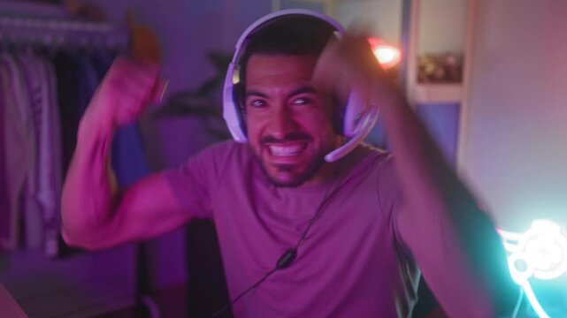 Man wearing white gaming headset clenches fist, shouts and smiles while seated at a neon lit gaming setup in studio; victory joy.