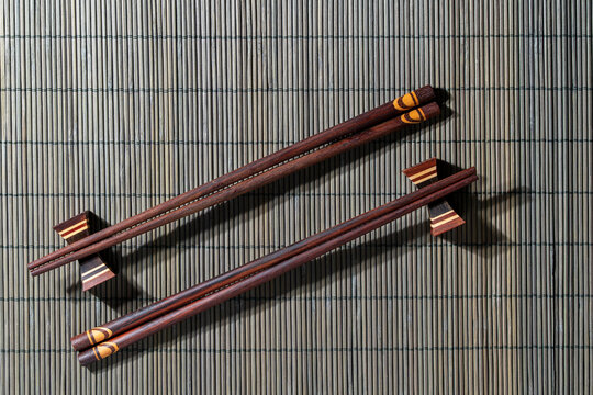 Wooden chopsticks on bamboo mat top view, closeup. Two pairs of dark wooden chopsticks with patterned tips resting on holders over grey bamboo rolling mat