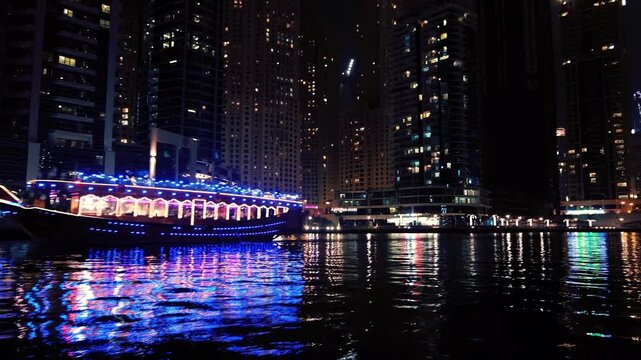Illuminated traditional dhow boat cruising along dubai marina at night with glowing skyscrapers and city lights in background.