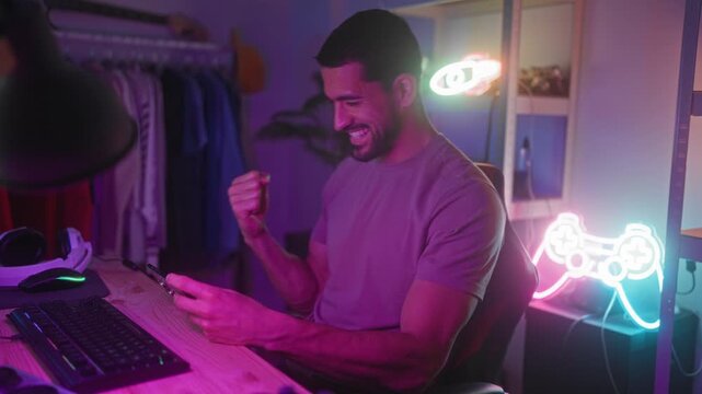 Man smiling and holding smartphone with both hands in a gaming studio with neon controller sign and keyboard visible; playful joy.
