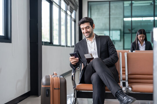 young male executive in suit holding passport with boarding pass, sitting in modern airport terminal checking time on a smartphone,preparing for corporate business trip or international flight