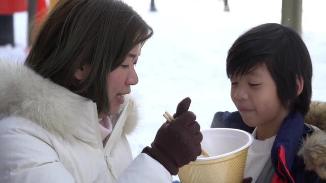 Asian mother feeding hot oden shirataki noodles to her son outdoors in winter.