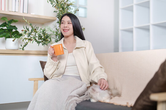 Asian woman drinking coffee while petting her cat on sofa at home