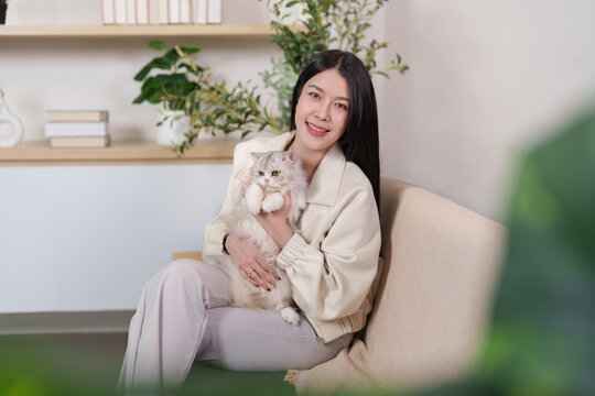 Asian woman holding cute cat while sitting on couch at home living room