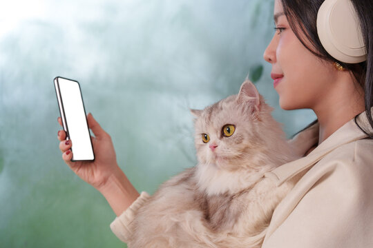 Asian woman wearing headphones holding fluffy cat while using smartphone with screen space