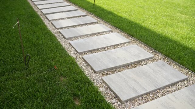 Concrete stepping stones path through green lawn with gravel edges. Rectangular concrete paving slabs laid diagonally on bright green grass edged with light-colored gravel. Landscape design, crushed s