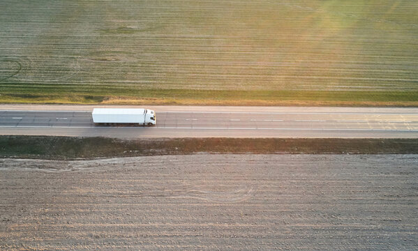 Aerial view of a white semi-truck driving along a highway through vast agricultural fields during golden hour in Mogilev, Mogilev Region, Belarus.