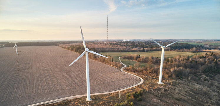 Aerial view of wind turbines standing in a rural landscape with plowed fields, a winding dirt road, and a distant radio tower under a soft sky in Mogilev, Mogilev Region, Belarus.