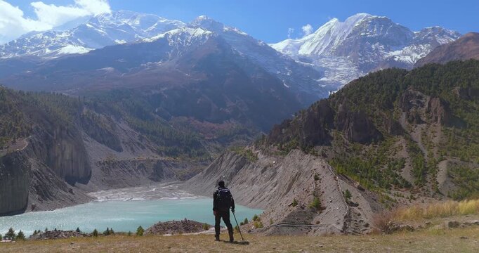 Annapurna Circuit, Manang, Nepal, drone glides toward a male hiker looking out over Gangapurna Lake and snow-capped mountain peaks on a bright sunny day