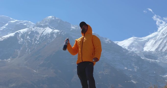 Annapurna Circuit, Manang Nepal, orbiting shot of a male hiker in a yellow jacket drinking water and enjoying the view against snow-capped Himalayan mountains
