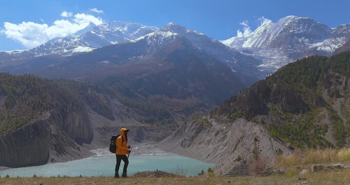 Annapurna Circuit, Manang, Nepal, drone moves towards a male hiker walking across a mountain trail overlooking Gangapurna Lake and Himalayan peaks.