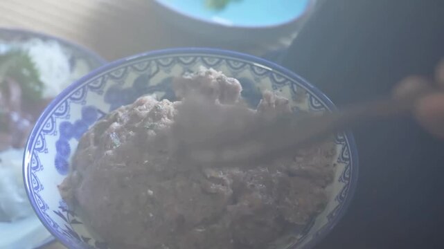 A person adds portions of minced meat paste into a pot of boiling vegetable broth using a wooden spoon and chopsticks, while other prepared dishes sit on the table