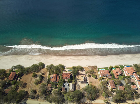 Aerial view of Playa Amarilla with beachfront houses, lush trees, and turquoise ocean waves crashing on the sandy shore in Tola, Rivas, Nicaragua.