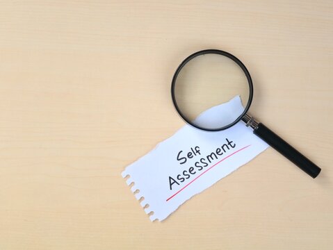 Self Assessment handwritten on ripped paper with a magnifying glass on a wooden desk background. Concept for personal development, performance review, audit, business strategy, and self-improvement