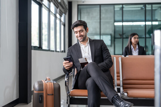 young male executive in suit holding passport with boarding pass, sitting in modern airport terminal checking time on a smartphone,preparing for corporate business trip or international flight