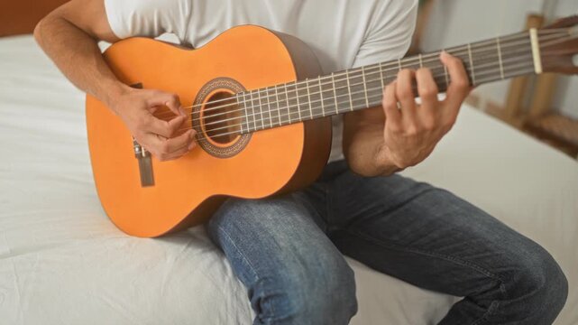 Man playing nylon string acoustic guitar, fingers fretting chords while seated on a bed in a bedroom; quiet practice serenity.