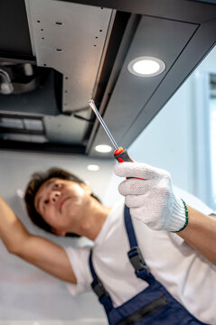 Asian male technician using screwdriver fixing extractor motor in cooker hood in modern kitchen. Maintenance worker man repairing exhaust fan in kitchen hood. Home appliance service on site