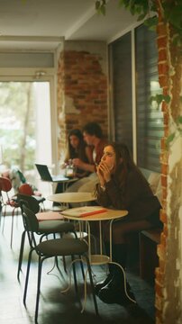 A young student is seen focused on her studies, writing in a notebook at a cafe table. Other students are visible in the background, engaged in various academic and social activities.
