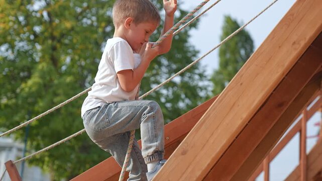 Child climbing rope playground. Child enjoys fun play outdoors. Child pulling rope developing strength coordination. Playful child at playground having fun climbing. Happy childhood outdoor adventure.