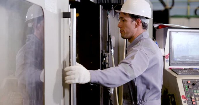 Asian man engineer closes CNC machine door after workspace review, completing equipment check and safety procedure before production cycle continues in precision manufacturing plant with compliance
